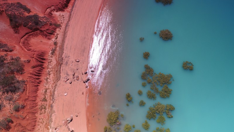 Aerial view of red sandy coastline meeting turquoise water with scattered green bushes.