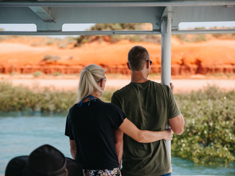 Couple standing on a boat, embracing, with a view of red cliffs and vegetation in the background.