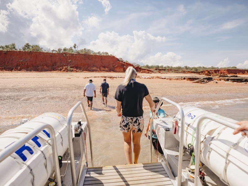 People disembarking a boat onto a sandy beach with red cliffs in the background.