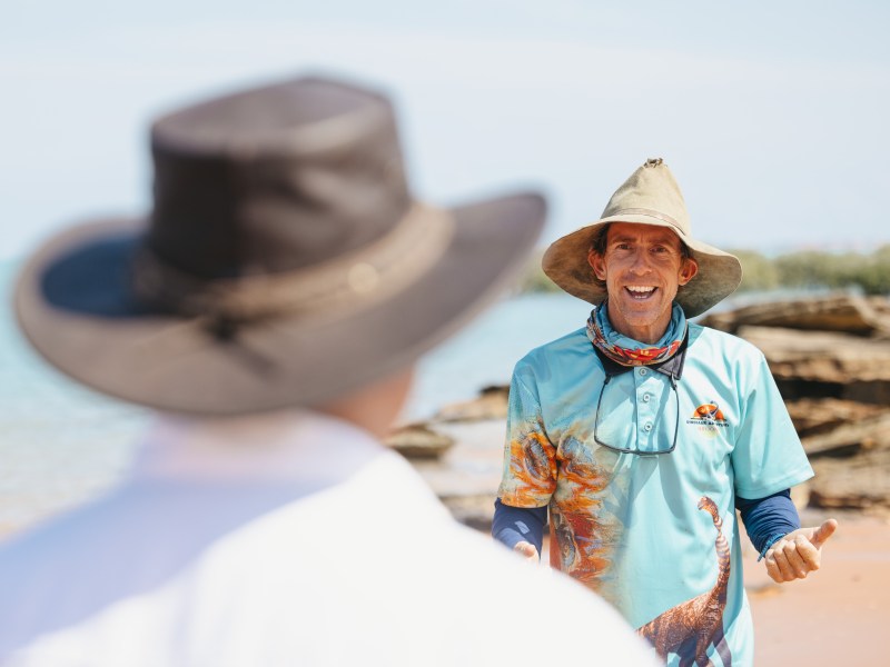 Man in hat and colorful shirt talks to another person on a rocky beach.
