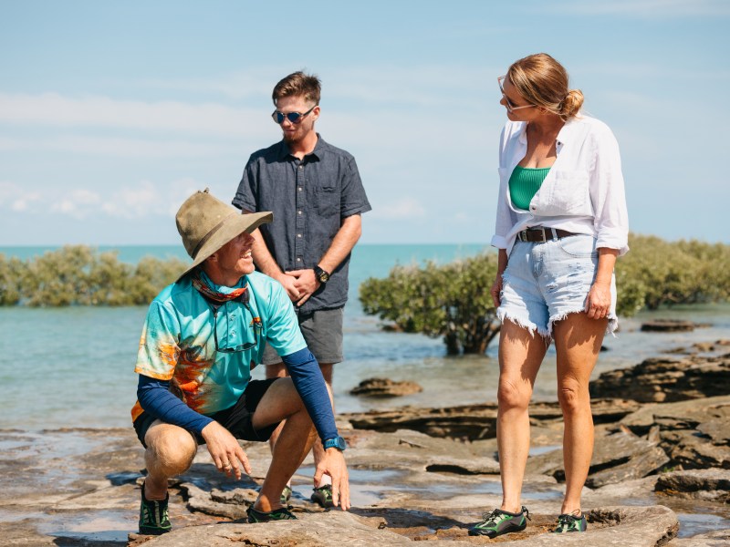 Three people on rocky shore with mangroves and ocean in background, one crouching, two standing.
