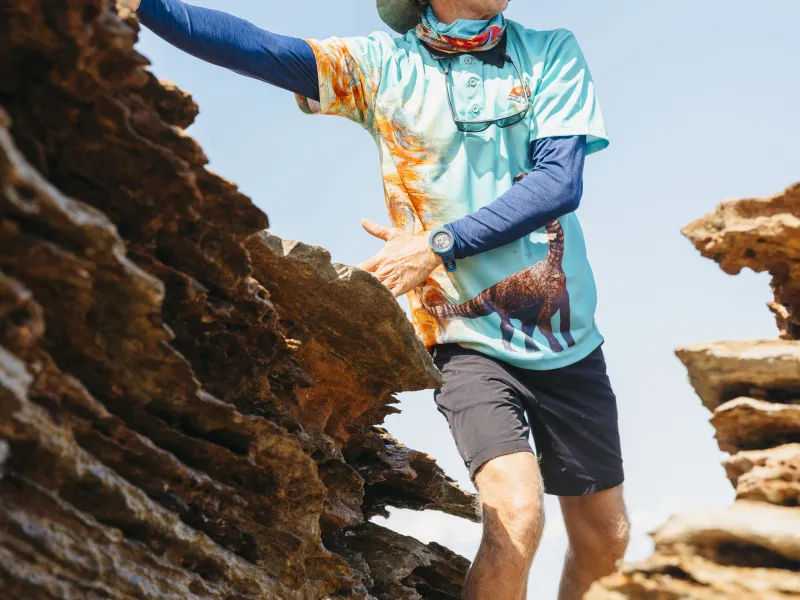 Man in a wide-brim hat and colorful shirt stands between rocks on a sunny day.