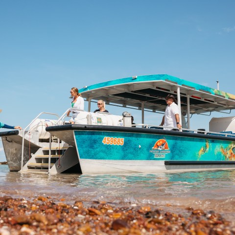 People boarding a colorful boat on a pebble beach under a clear sky.
