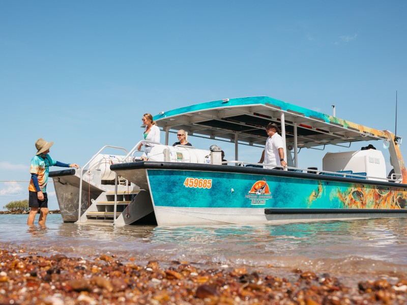 People boarding a colorful boat on a pebble beach under a clear sky.
