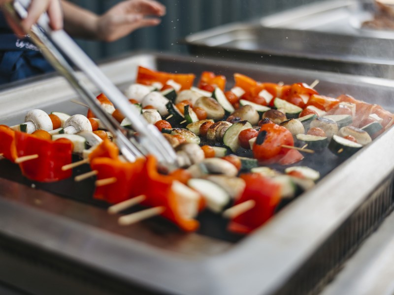 Person grilling vegetable skewers, including peppers and mushrooms, on a flat top grill.