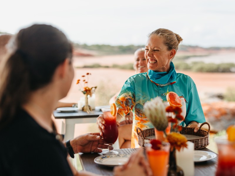Smiling woman serving drinks at an outdoor setting with a scenic background.