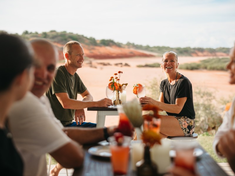 Group of people smiling and having drinks at an outdoor table with desert landscape in the background.