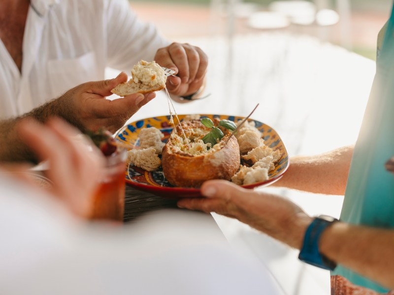 Hands holding a bread bowl with dip and torn bread pieces on a colorful plate.
