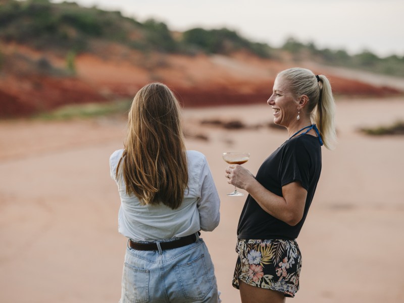 Two women smiling outdoors, one holding a drink, with sandy landscape in background.