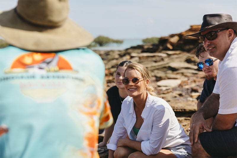 Group of people sitting outdoors, some wearing hats and sunglasses, with rocky terrain in the background.